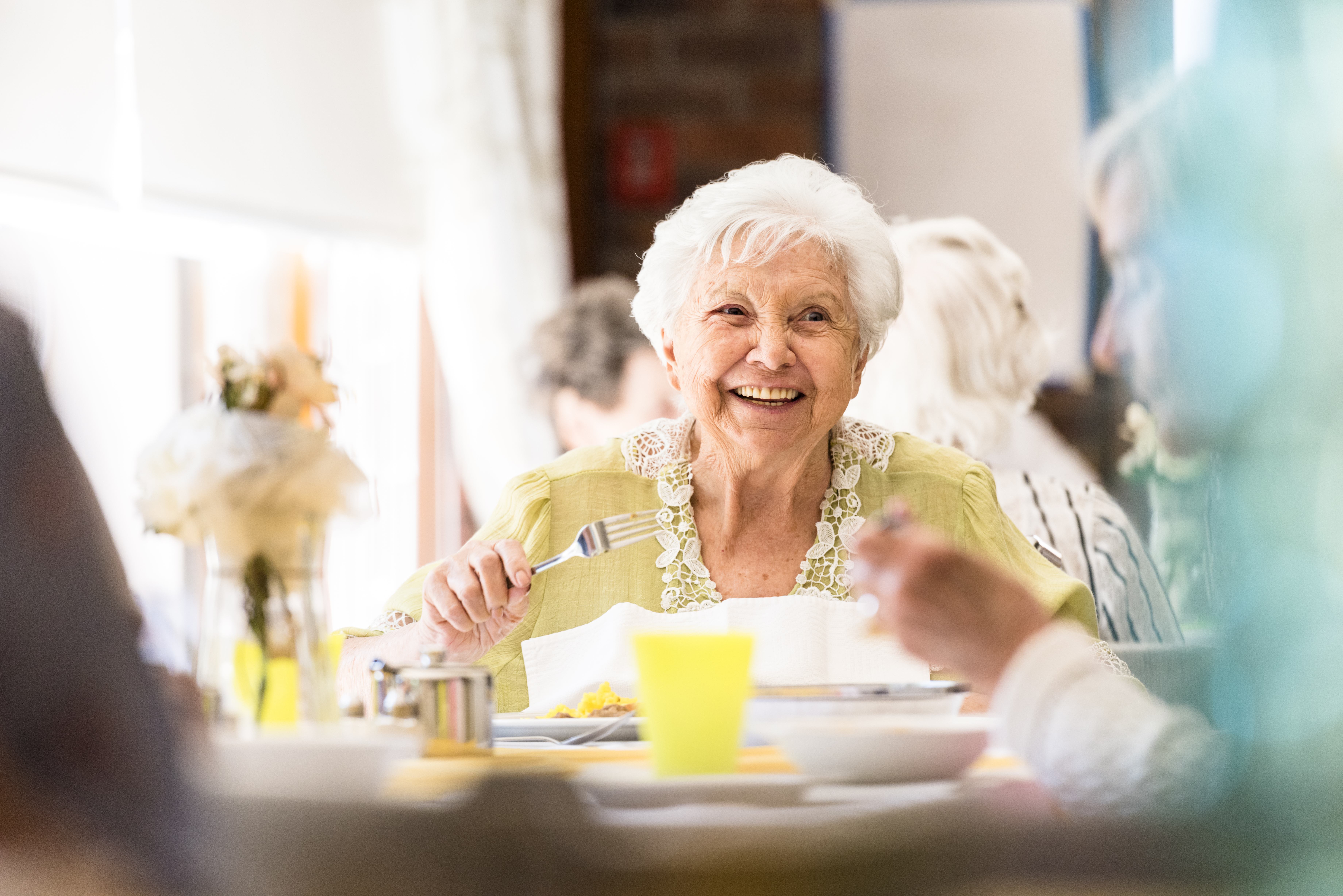 Äldre dam med vitt hår skrattar. Hon håller i en sked och framför henne står en skål. Hon har en gul blus på sig med blommor.
